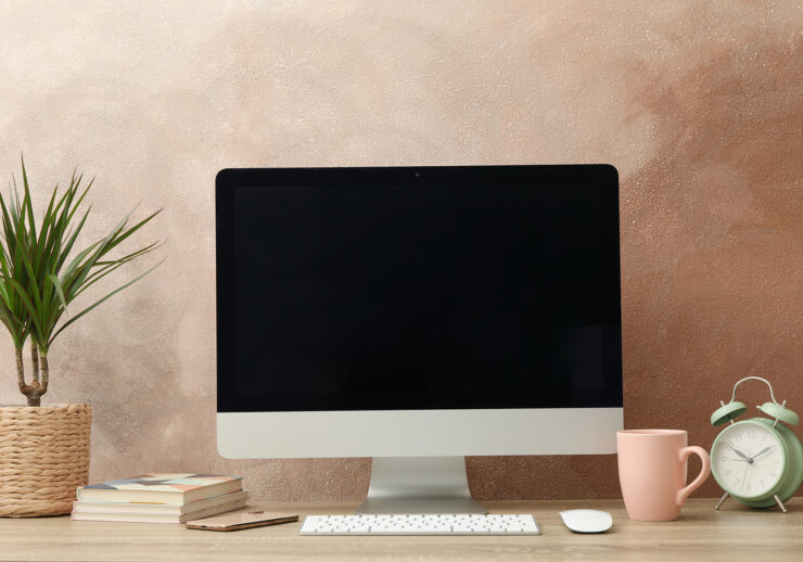 Workplace with computer, plant and alarm clock on wooden table.